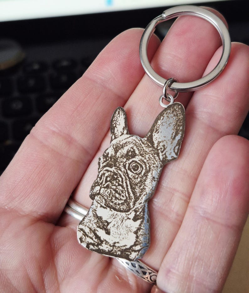 May include: A silver-toned metal keyring featuring a laser-engraved portrait of a French Bulldog. The charm is shaped to the outline of the dog's head and ears, attached to a circular split ring. The engraving displays dark, detailed shading.