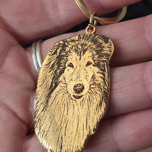 May include: A gold-toned metal keyring featuring a laser-engraved portrait of a long-haired collie dog. The pendant has an irregular, organic shape that follows the outline of the dog's head and fur, attached to a circular gold-coloured split ring.