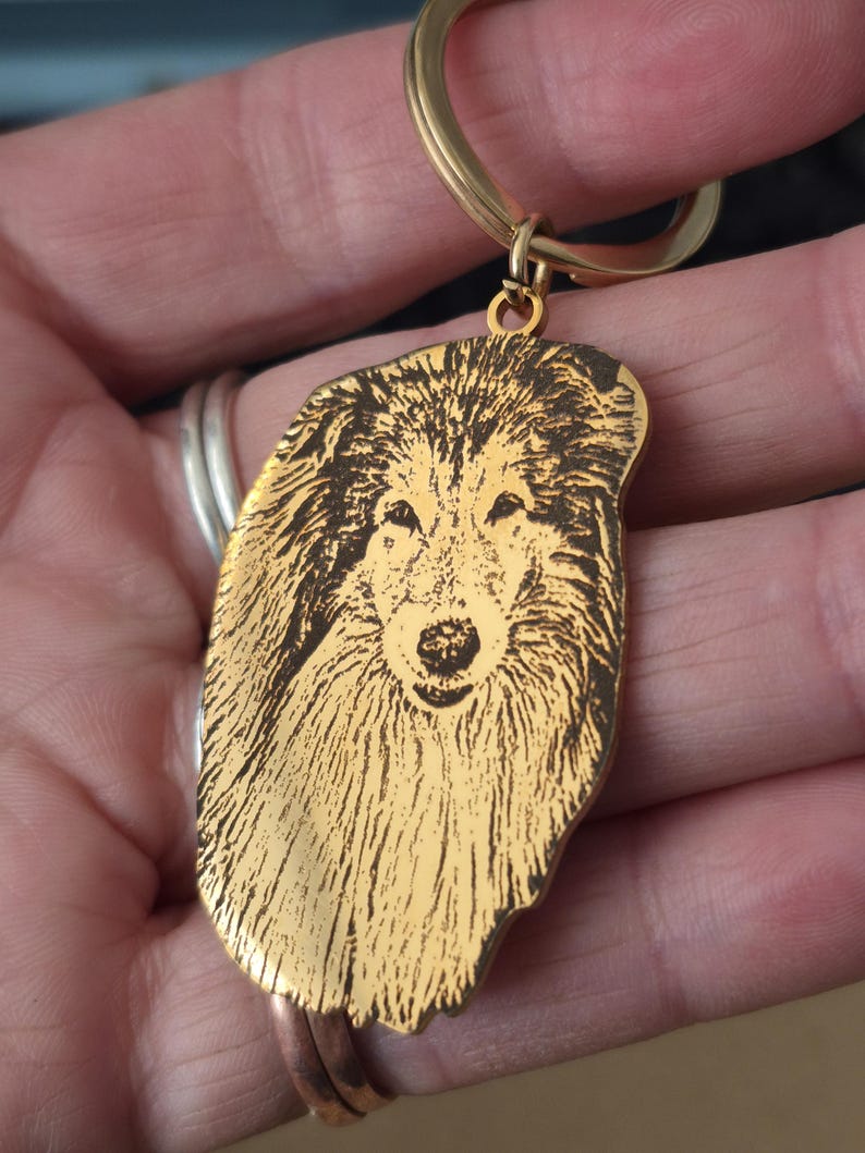 May include: A gold-toned metal keyring featuring a laser-engraved portrait of a long-haired collie dog. The pendant has an irregular, organic shape that follows the outline of the dog's head and fur, attached to a circular gold-coloured split ring.