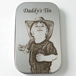Puede incluir: Una lata ovalada de color plateado con las palabras "Daddy's Tin" grabadas en la parte superior. La lata presenta una imagen grabada de un niño con sombrero de vaquero y camiseta. El niño sonríe y mira hacia arriba.