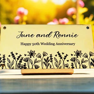 May include: A rectangular, gold-colored plaque with the names "June and Ronnie" and the text "Happy 50th Wedding Anniversary." The bottom edge features a black floral design. The plaque is mounted on a wooden stand.