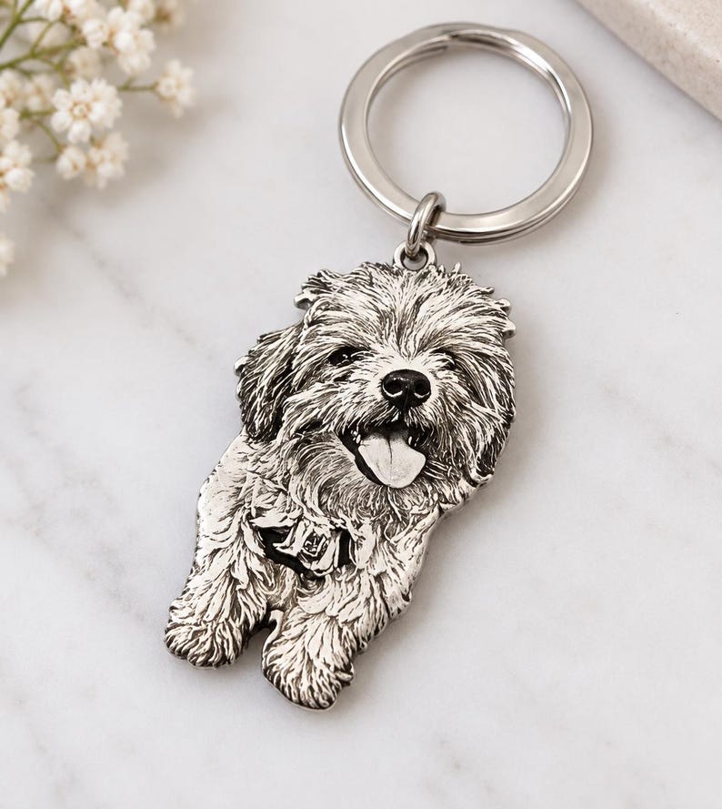 May include: A silver-toned metal keyring featuring a detailed, embossed portrait of a fluffy dog with its tongue out. The pendant is attached to a circular split ring, showcasing intricate textures that mimic the appearance of thick fur.