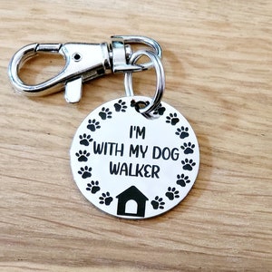 Puede incluir: Llavero de plata con una etiqueta redonda que dice "Estoy con mi paseador de perros" rodeado de huellas de patas y un icono de casa.