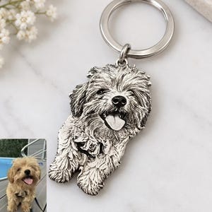 May include: A silver-toned metal keyring featuring a detailed, engraved portrait of a fluffy dog with its tongue out. The charm is attached to a circular split ring. A small inset photograph shows the original dog with light brown fur.