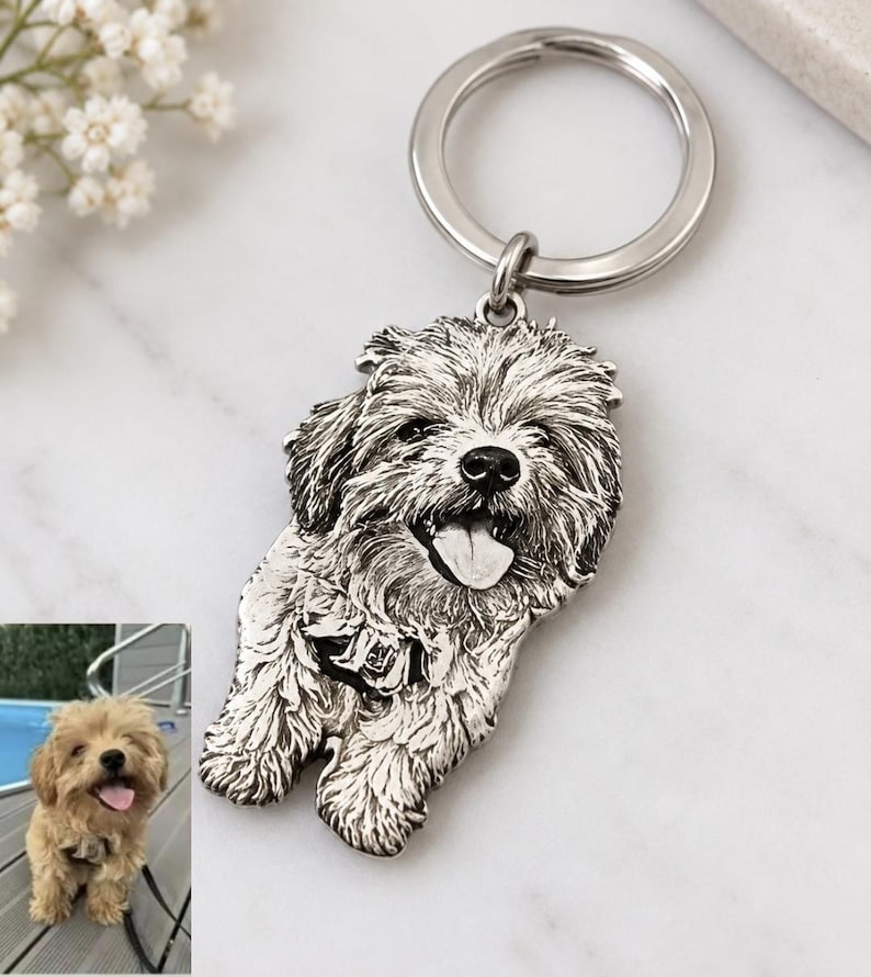 May include: A silver-toned metal keyring featuring a detailed, engraved portrait of a fluffy dog with its tongue out. The charm is attached to a circular split ring. A small inset photograph shows the original dog with light brown fur.
