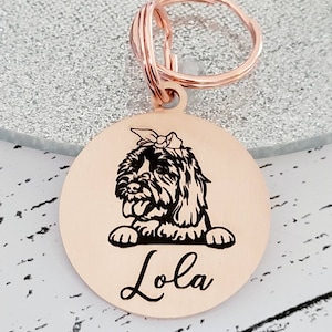 May include: Rose gold dog tag featuring a black and white silhouette of a dog with a bow on its head. The dog tag is engraved with the name "Lola".