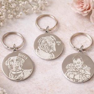 May include: Three silver-colored keychains with engraved portraits. One keychain features a dog named "Betty", another a dog named "Roo", and the third a child holding a puppy. Each keychain has a silver ring.
