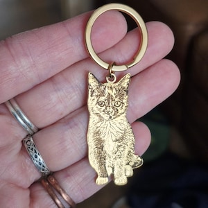May include: A gold-toned metal keyring featuring a flat, laser-engraved charm in the shape of a sitting kitten. The charm displays a detailed, dark-etched portrait of a cat on a polished gold surface, attached to a circular gold split ring.