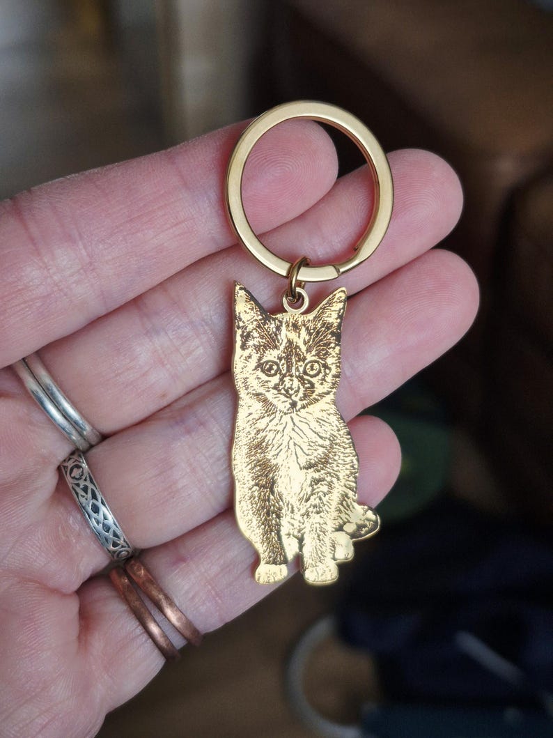May include: A gold-toned metal keyring featuring a flat, laser-engraved charm in the shape of a sitting kitten. The charm displays a detailed, dark-etched portrait of a cat on a polished gold surface, attached to a circular gold split ring.