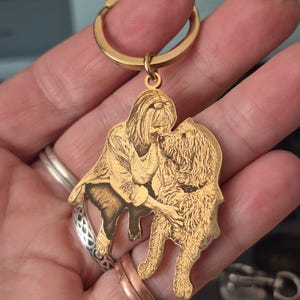 May include: A gold-toned metal keyring featuring a custom laser-engraved illustration of a person kneeling and interacting with a long-haired dog. The pendant is shaped to follow the outline of the figures and includes a circular split ring attachment.
