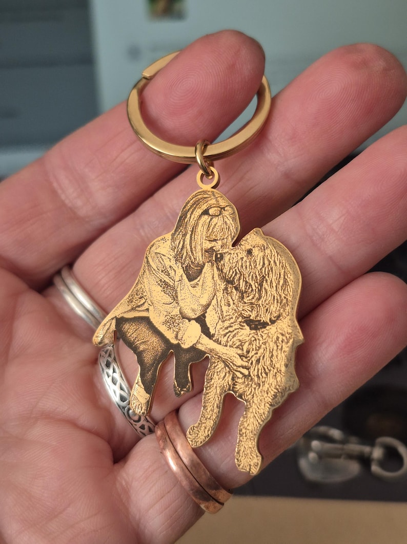 May include: A gold-toned metal keyring featuring a custom laser-engraved illustration of a person kneeling and interacting with a long-haired dog. The pendant is shaped to follow the outline of the figures and includes a circular split ring attachment.