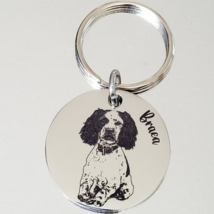 May include: Silver dog tag keychain with a black and white photo of a dog engraved on it. The dog's name, "Braea", is engraved on the tag.