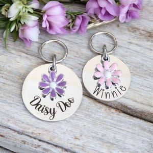 Puede incluir: Dos chapas de identificación para perros de plata con un dije de flor. La chapa de la izquierda dice "Daisy Doo" y la de la derecha dice "Winnie".