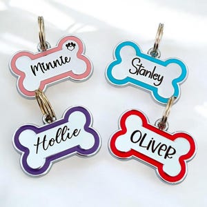 Könnte beinhalten: Vier silberne, knochenförmige Hundemarken mit bunten Emaille-Rändern. Die Marken sind mit den Namen "Minnie", "Stanley", "Hollie" und "Oliver" graviert.