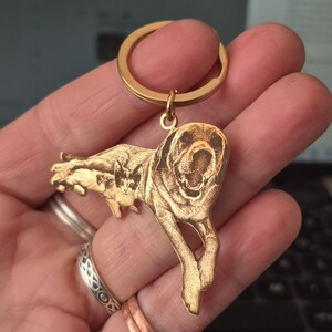 May include: A gold-toned metal keyring featuring a custom laser-engraved illustration of a dog and a cat. The keyring has a circular split ring at the top and a flat, shaped pendant depicting the two animals in a detailed, etched style.