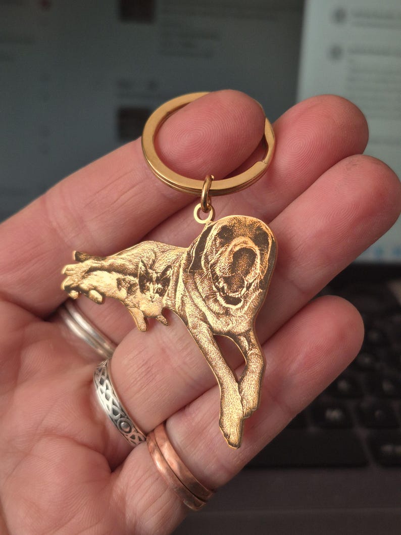 May include: A gold-toned metal keyring featuring a custom laser-engraved illustration of a dog and a cat. The keyring has a circular split ring at the top and a flat, shaped pendant depicting the two animals in a detailed, etched style.
