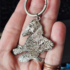 May include: A silver-toned metal keyring featuring a detailed, laser-engraved portrait of a dog with long, wavy fur. The dog is depicted in a seated position, and the metal charm is attached to a circular split ring for keys.