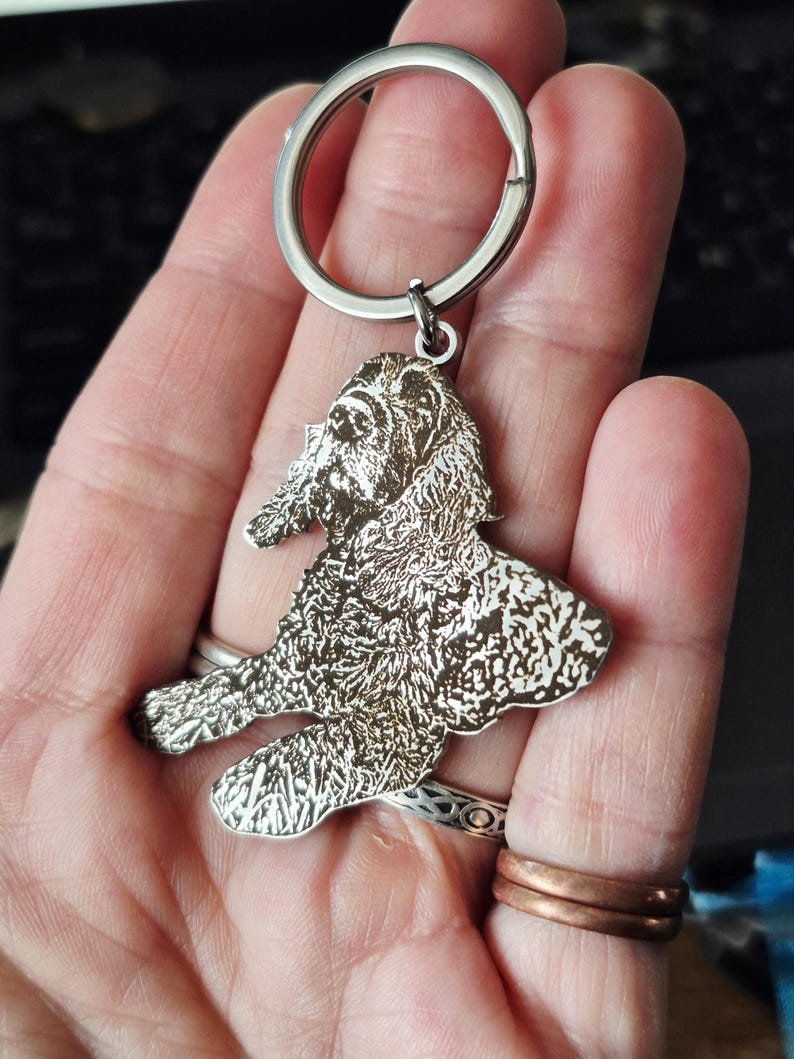 May include: A silver-toned metal keyring featuring a detailed, laser-engraved portrait of a dog with long, wavy fur. The dog is depicted in a seated position, and the metal charm is attached to a circular split ring for keys.