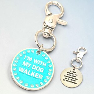 Peut inclure: Deux porte-clés pour chien. L'un est un cercle turquoise avec le texte "I'M WITH MY DOG WALKER" et des empreintes de pattes. L'autre est un cercle plus petit, de couleur dorée, avec les coordonnées : "IF LOST PLEASE CALL DOG TIRED ON 01234567890 OR MY MUM ON 01234567890". Les deux ont des fermoirs argentés.