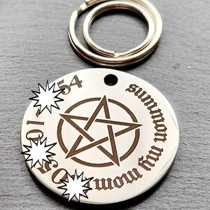May include: A silver-toned metal keychain with a round tag featuring a pentagram and the text "summon thy demons" in a gothic font. The tag has a hole at the top for attaching to a keyring.