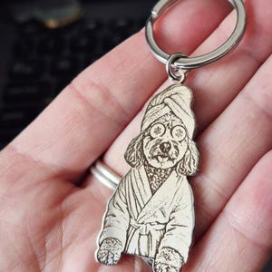 May include: A silver-toned metal keyring featuring a laser-engraved illustration of a dog wearing a bathrobe, a towel wrapped around its head, and cucumber slices over its eyes. The charm is attached to a circular metal split ring.