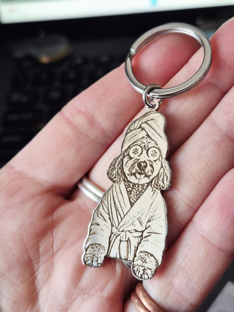 May include: A silver-toned metal keyring featuring a laser-engraved illustration of a dog wearing a bathrobe, a towel wrapped around its head, and cucumber slices over its eyes. The charm is attached to a circular metal split ring.