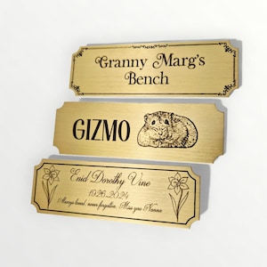 May include: Three gold-colored rectangular plaques with engraved text and designs. The top plaque reads "Granny Marg's Bench." The middle plaque says "GIZMO" with a hamster illustration. The bottom plaque reads "Enid Dorothy Vine 1926-2024."
