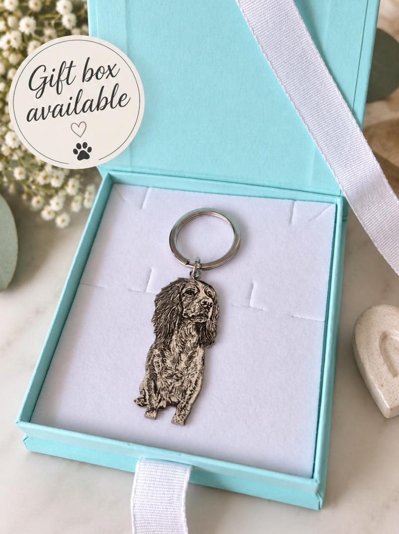 May include: A silver-toned metal keyring featuring a detailed engraved portrait of a long-eared dog, presented inside a light blue gift box with a white ribbon. A circular sticker on the box lid displays the text Gift box available with a heart and paw print.