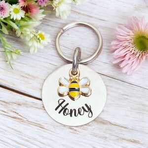 May include: A silver round pet tag with a bee charm and the name "Honey" engraved in black.