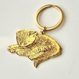 May include: A gold-coloured metal keyring featuring a flat, laser-engraved charm in the shape of a sleeping cat. The charm displays a detailed, textured illustration of a feline curled up with its eyes closed, attached to a circular split ring.