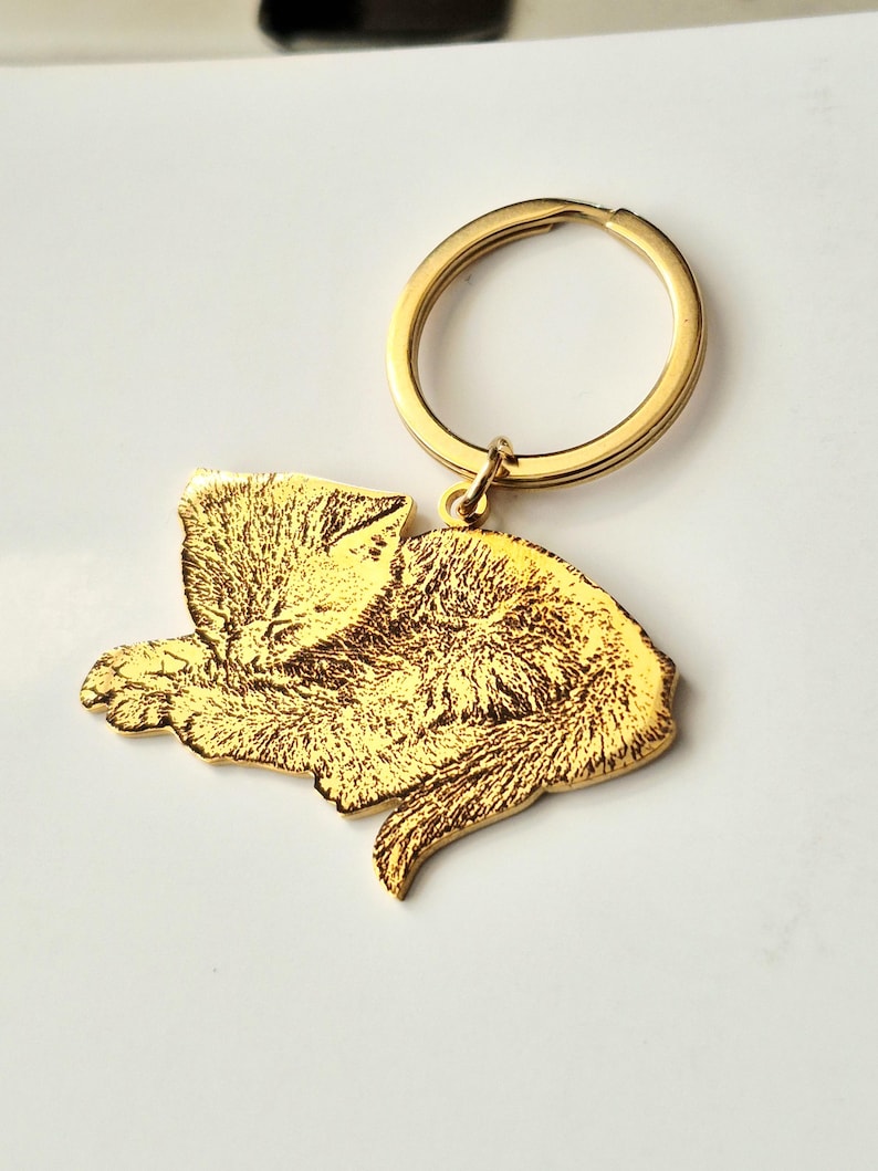 May include: A gold-coloured metal keyring featuring a flat, laser-engraved charm in the shape of a sleeping cat. The charm displays a detailed, textured illustration of a feline curled up with its eyes closed, attached to a circular split ring.