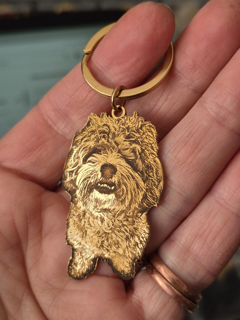 May include: A gold-toned metal keyring featuring a custom laser-engraved portrait of a shaggy-haired dog. The pendant is shaped to the outline of the dog's head and upper body, attached to a circular split ring for keys.