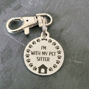 May include: Silver dog tag with a black engraved design. The tag reads "I'm with my pet sitter" and features a bone and paw prints around the edge. The tag is attached to a silver split ring and a silver clip.
