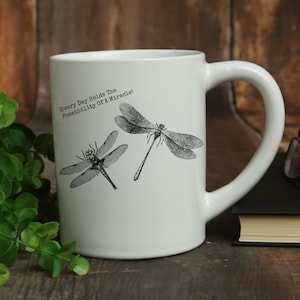 May include: White ceramic mug with a black and white dragonfly design. The mug has a handle and the text "Every Day Holds The Possibility Of A Miracle!" printed on the front.