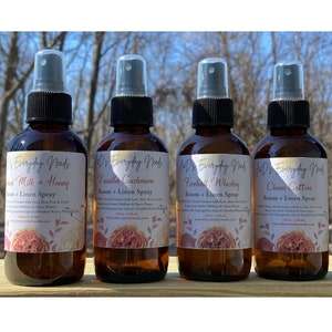 May include: Four brown glass spray bottles with black spray tops and white labels. The labels have the text "JD's Everyday Needs" and the scent names "Vanilla Cashmere", "Fireball Whiskey", "Clean Cotton", and "Cinnamon Milk & Honey". Each bottle has the text "Room + Linen Spray" on the label.