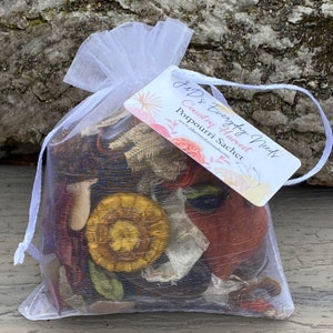 May include: A white mesh bag filled with dried flowers and leaves. The bag is tied with a white ribbon and has a tag attached that reads "J & D's Everyday Needs Country Harvest Potpourri Sachet www.j&dseverydayneeds.com".