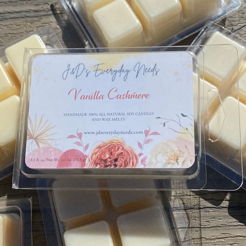 May include: A clear plastic container with four soy wax melts in a grid pattern. The wax melts are a light yellow color. The container has a label that reads "J&D's Everyday Needs" and "Vanilla Cashmere". The label also states that the wax melts are handmade with 100% all natural soy candles and wax melts. The label includes the website address "www.jdseverydayneeds.com". The label has a floral design with pink, peach, and yellow colors.