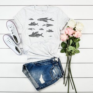 May include: A white t-shirt with a black and white illustration of flying fish. The shirt is laid out on a white wooden surface with a pair of white sneakers, denim shorts, and a bouquet of pink and white roses.