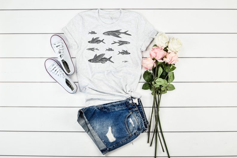 May include: A white t-shirt with a black and white illustration of flying fish. The shirt is laid out on a white wooden surface with a pair of white sneakers, denim shorts, and a bouquet of pink and white roses.