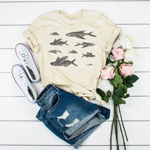 May include: A beige t-shirt with a black and white illustration of flying fish. The t-shirt is paired with blue denim shorts, white sneakers, and pink roses.