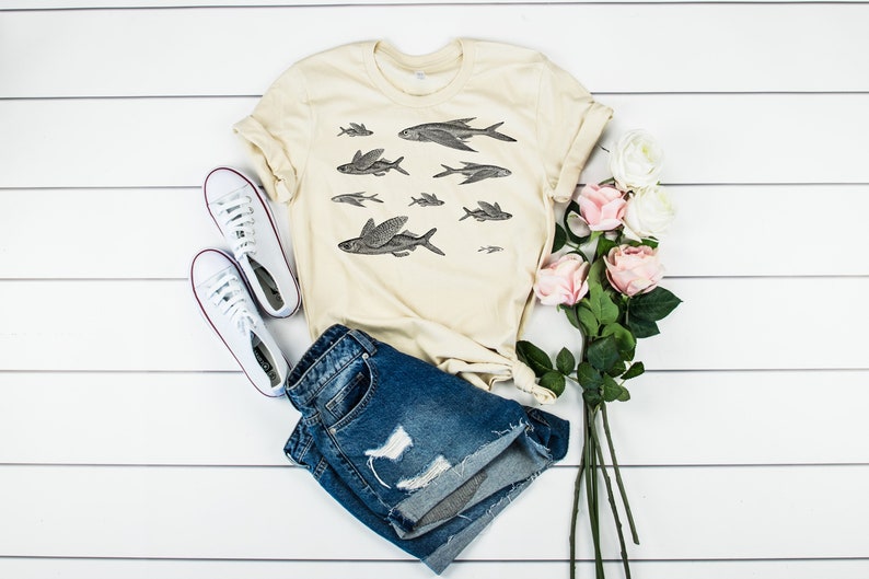 May include: A beige t-shirt with a black and white illustration of flying fish. The t-shirt is paired with blue denim shorts, white sneakers, and pink roses.