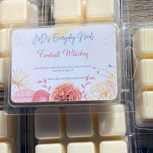 May include: A white wax melt with a floral design label that reads "J&D's Everyday Needs Fireball Whiskey Handmade 100% All Natural Soy Candles and Wax Melts www.jdseverydayneeds.com 3.1 fl. oz./Net Wt 2.7 oz. 176.5 g)"
