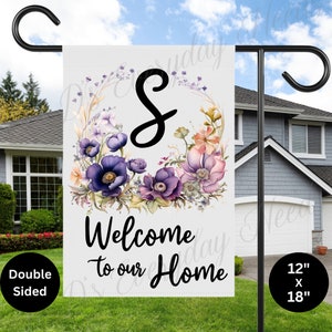 May include: A white welcome garden flag featuring a floral wreath with the letter "S" in the centre. The wreath is made of purple, pink and white flowers. The text "Welcome to our Home" is printed in black below the wreath. The flag measures 12 inches by 18 inches.