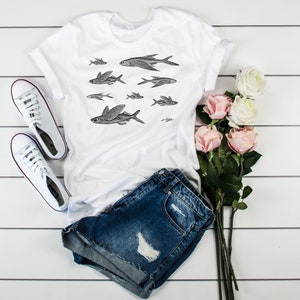 May include: White t-shirt with a black and white illustration of flying fish. The shirt is paired with denim shorts and white sneakers. There are also pink roses in the image.