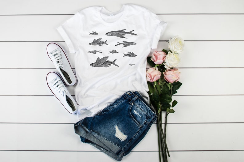 May include: White t-shirt with a black and white illustration of flying fish. The shirt is paired with denim shorts and white sneakers. There are also pink roses in the image.