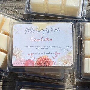 May include: A clear plastic container with six individual wax melts. The wax melts are a light yellow color. The container has a label that reads "J&D's Everyday Needs Clean Cotton Handmade 100% All Natural Soy Candles and Wax Melts www.jdseverydayneeds.com 3.1 fl. oz./Net Wt. 2.7 oz. (76.5 g)"