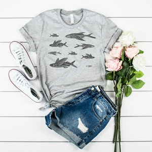 May include: A gray t-shirt with a graphic print of flying fish. The shirt is paired with denim shorts and white sneakers. A bouquet of pink and white roses is on the right side of the image.