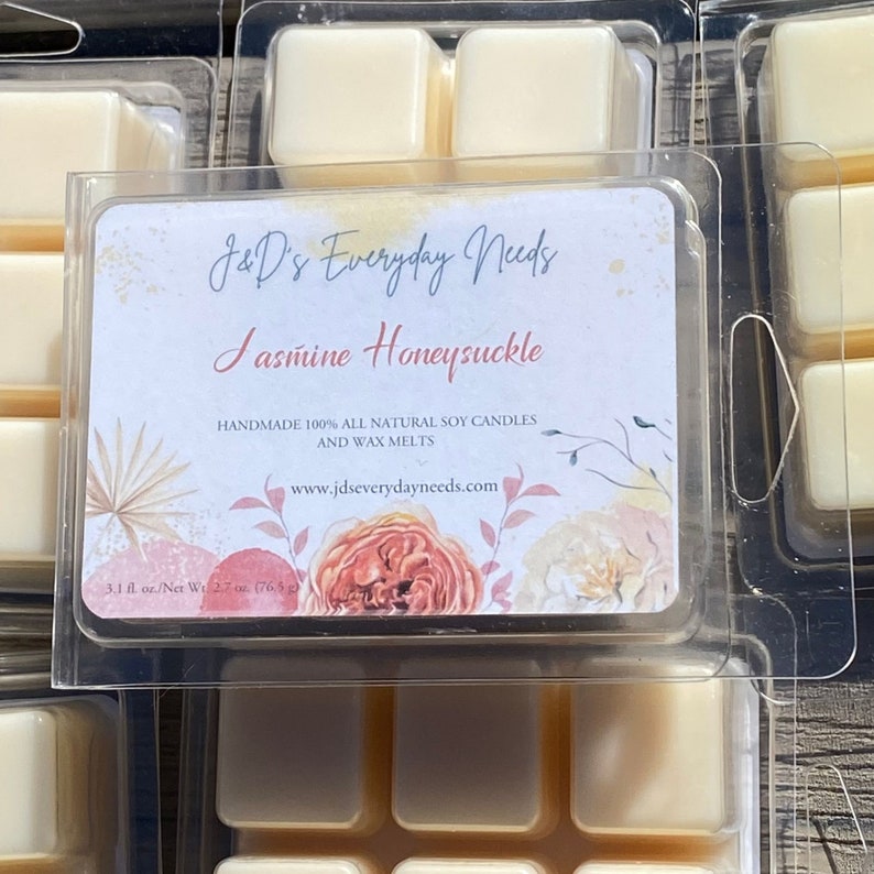 May include: A six-pack of soy wax melts in a clear plastic container. The label reads "J&D's Everyday Needs Jasmine Honeysuckle Handmade 100% All Natural Soy Candles and Wax Melts www.jdseverydayneeds.com 3.1 fl. oz./Net Wt. 2.7 oz. (76.5 g)"