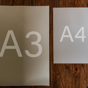 Op de afbeelding: Twee vellen papier, een met het label "A3" en het andere met het label "A4", liggen op een houten oppervlak. Het vel met het label "A3" is groter dan het vel met het label "A4".