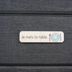 May include: A wooden sign with the text "Je mets la table" in black lettering. The sign also features a simple illustration of a plate, fork, and knife.